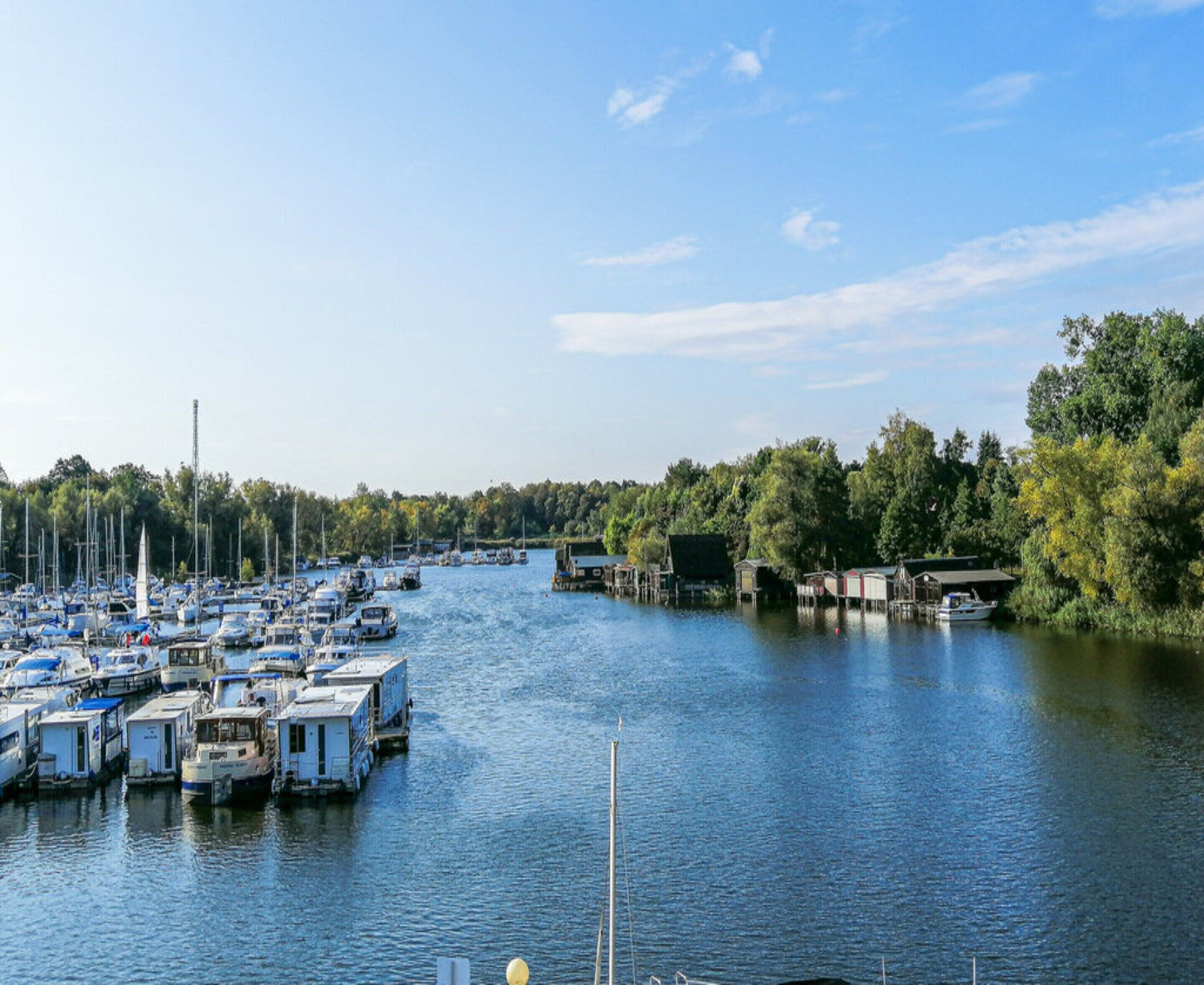A marina with numerous boats docked along a river, surrounded by trees and small buildings.