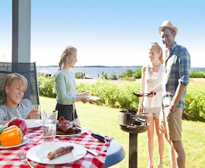 Une famille prend son repas à l’extérieur, près d’un barbecue, autour d’une table garnie de plats, avec une vue sur l’eau.