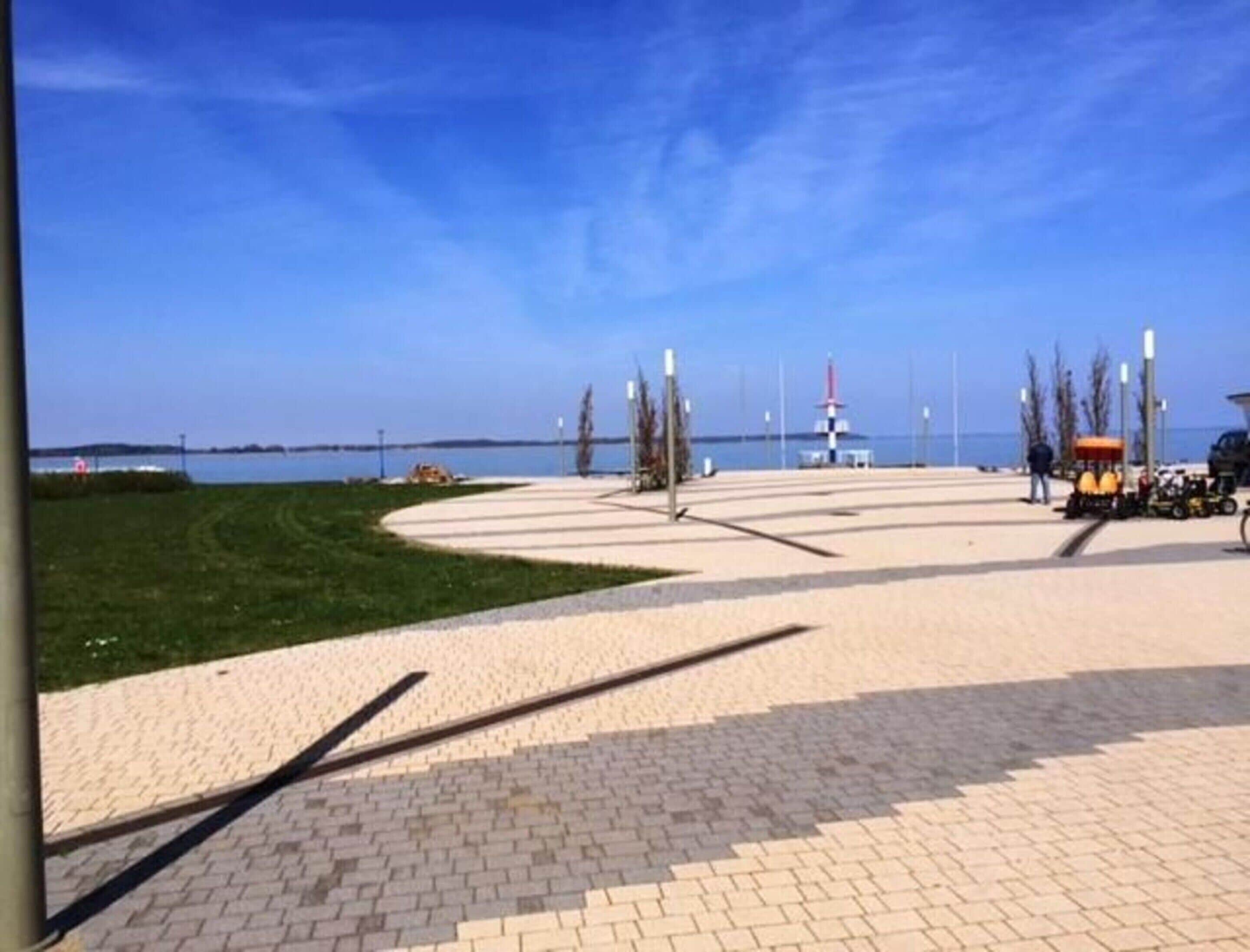 A paved waterfront promenade with a grassy area, a small kiosk, and a distant lighthouse.