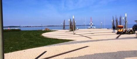A paved waterfront promenade with a grassy area, a small kiosk, and a distant lighthouse.