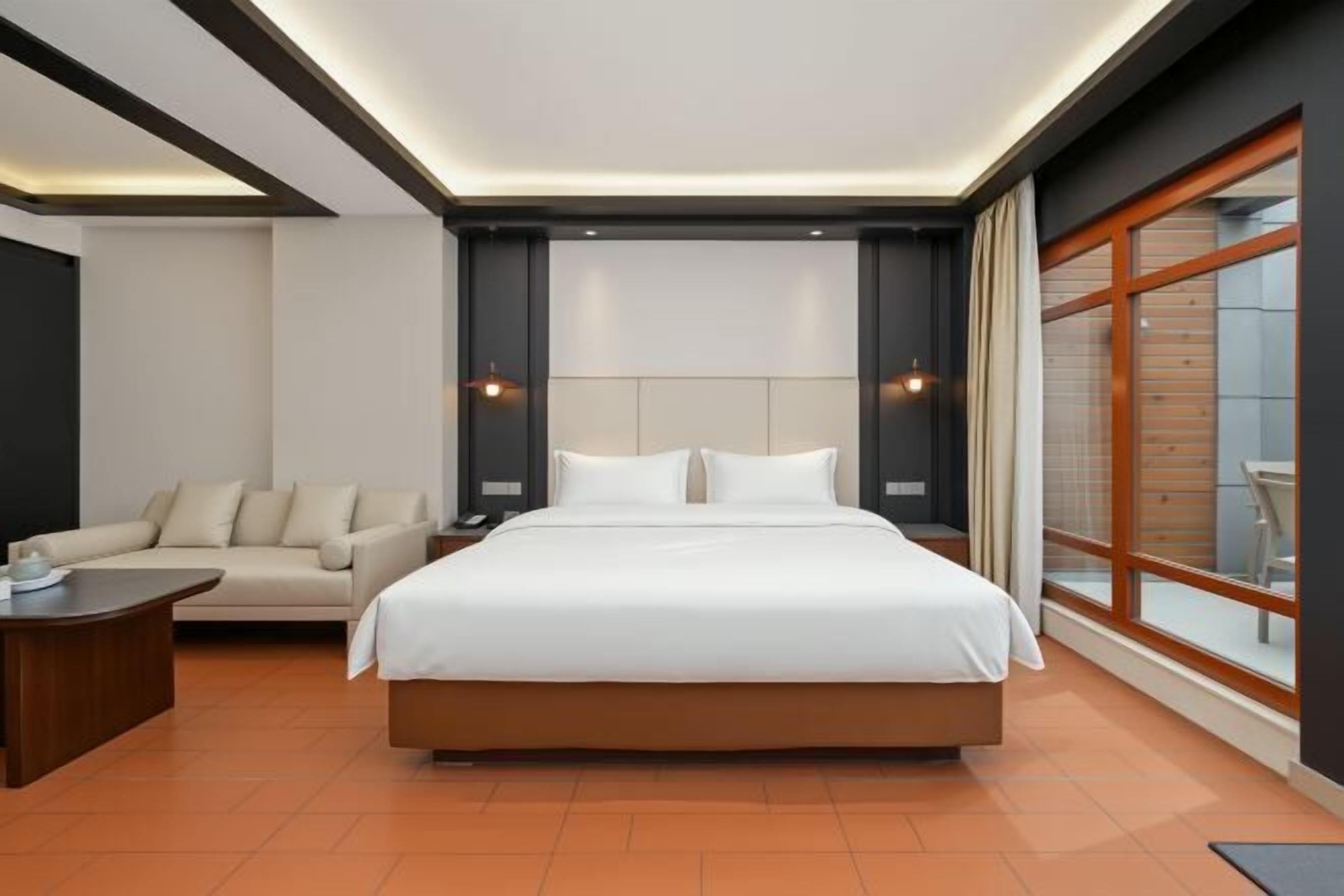 A modern hotel room with a large bed, a sofa, a nightstand, and a large window.