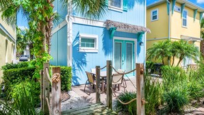 Um deck de madeira leva a uma casa azul com telhado de palha, rodeada por plantas tropicais e um céu claro.