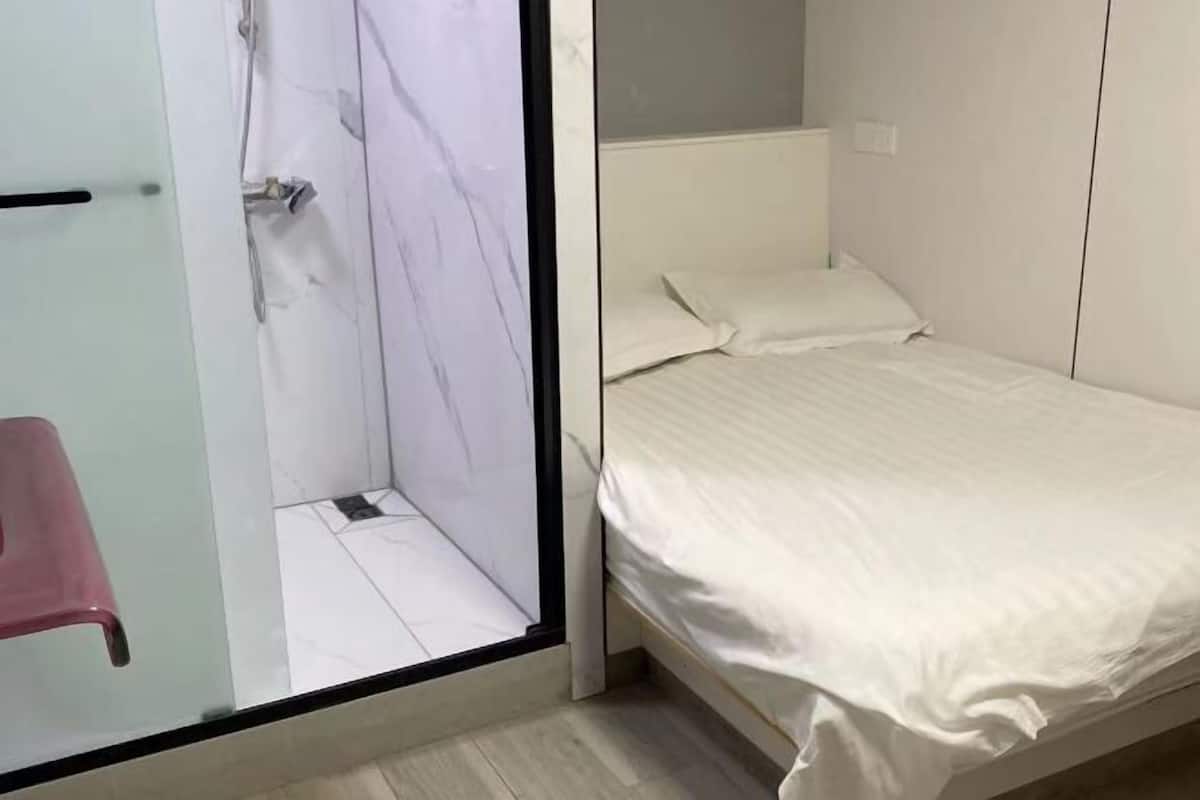 A bedroom with a bed, a small stool, and a shower area.