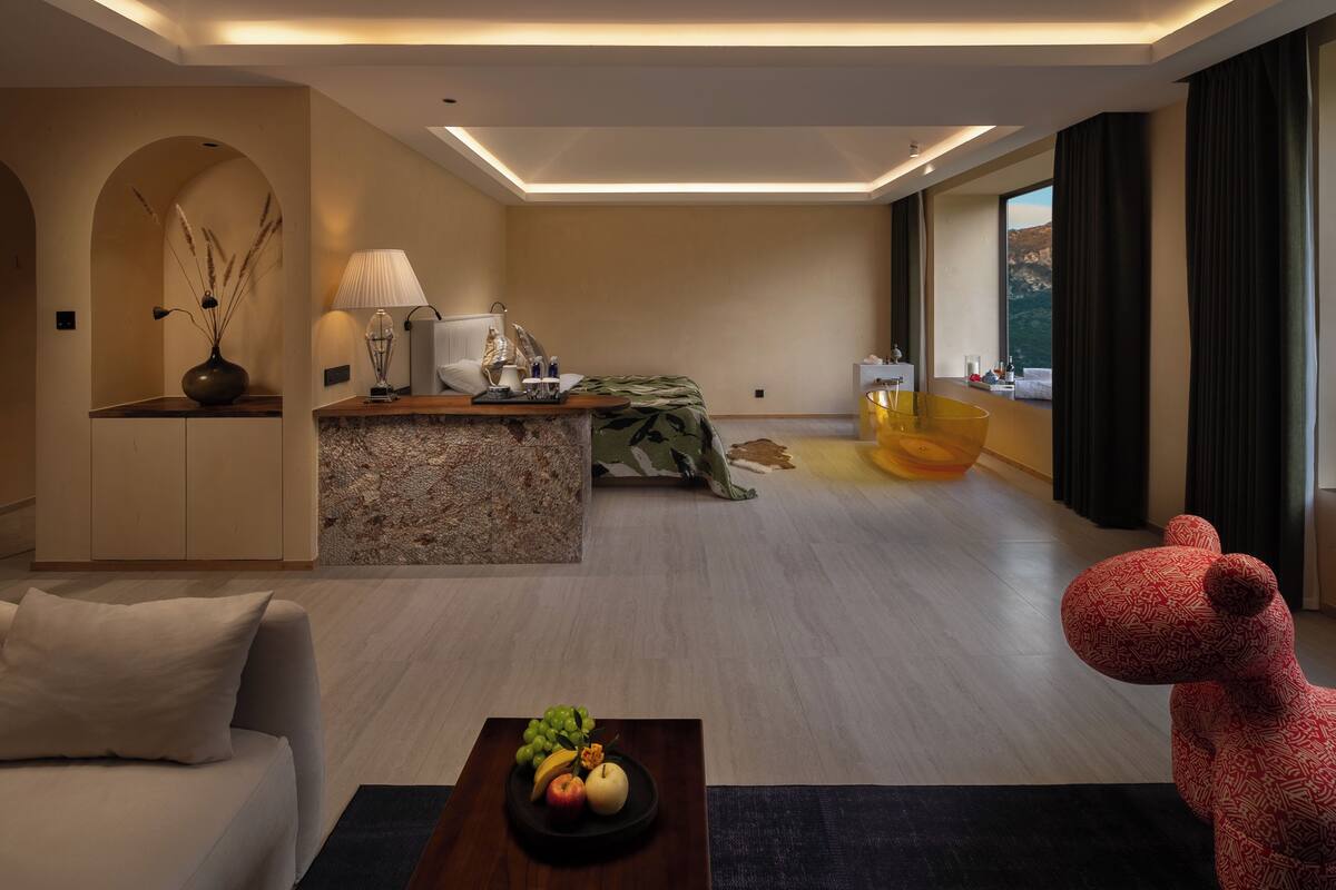 A modern living room with a sofa, a coffee table with fruit, a red bear sculpture, a kitchen area with a lamp, and a large window with curtains.