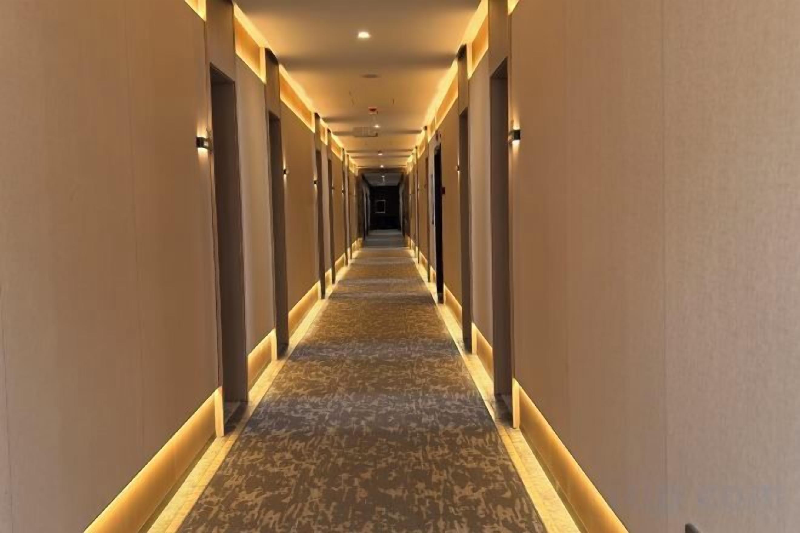 A long, narrow hotel corridor with carpeted floors and walls, illuminated by overhead lighting.