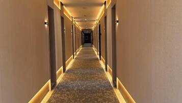 A long, narrow hotel corridor with carpeted floors and walls, illuminated by overhead lighting.