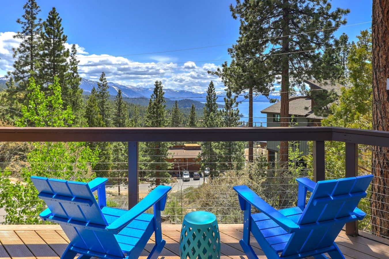 A balcony with two blue chairs and a small table, overlooking a mountain landscape and a parking area.