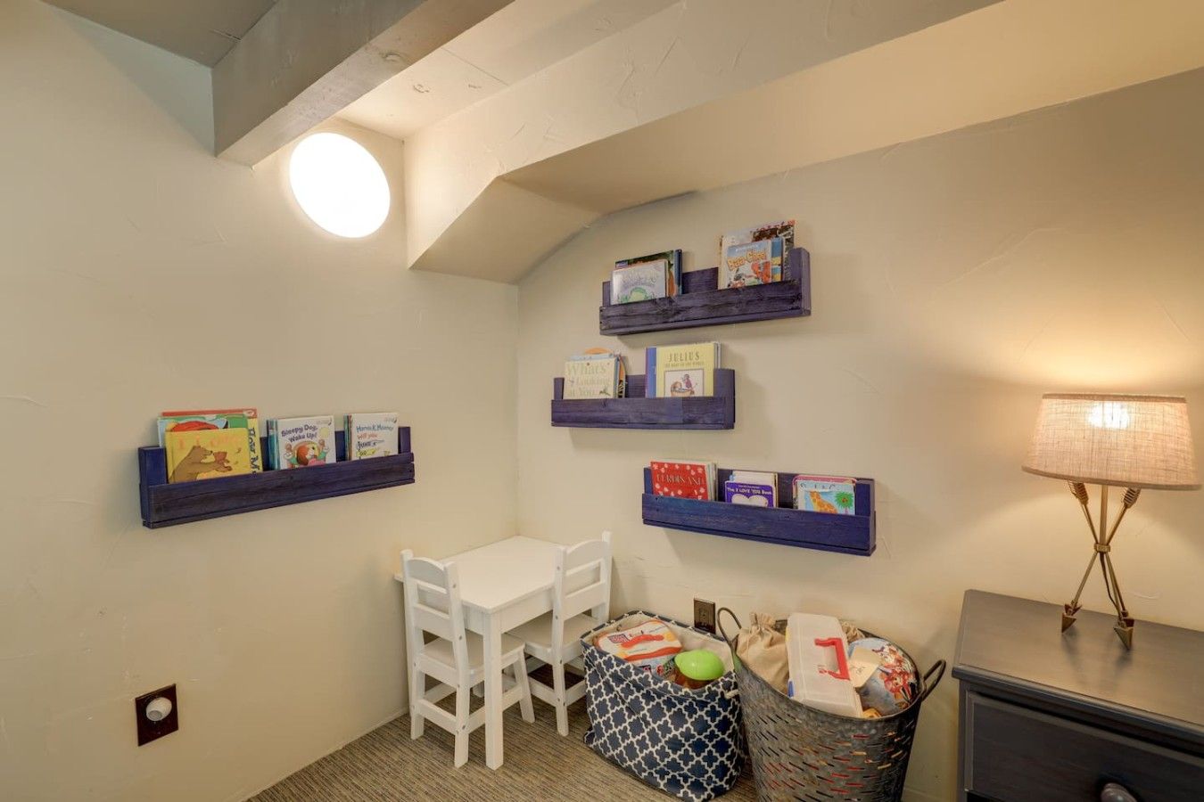 Ein kleines Zimmer mit einem weißen Tisch, einem Stuhl und einer Lampe. An den Wänden befinden sich Bücherregale mit Büchern.