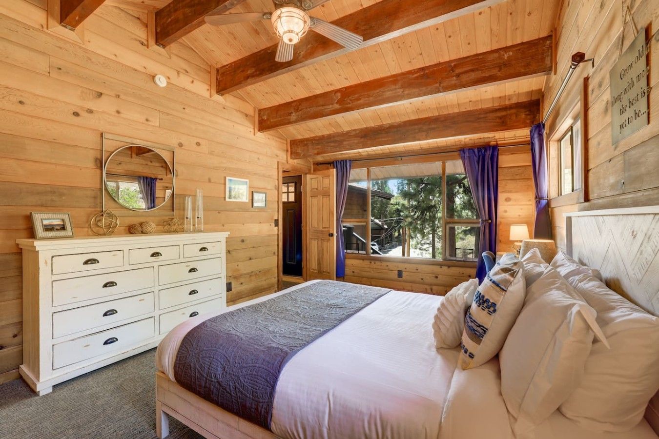 Un dormitorio de cabaña de madera con cama, cómoda y ventana con cortinas.