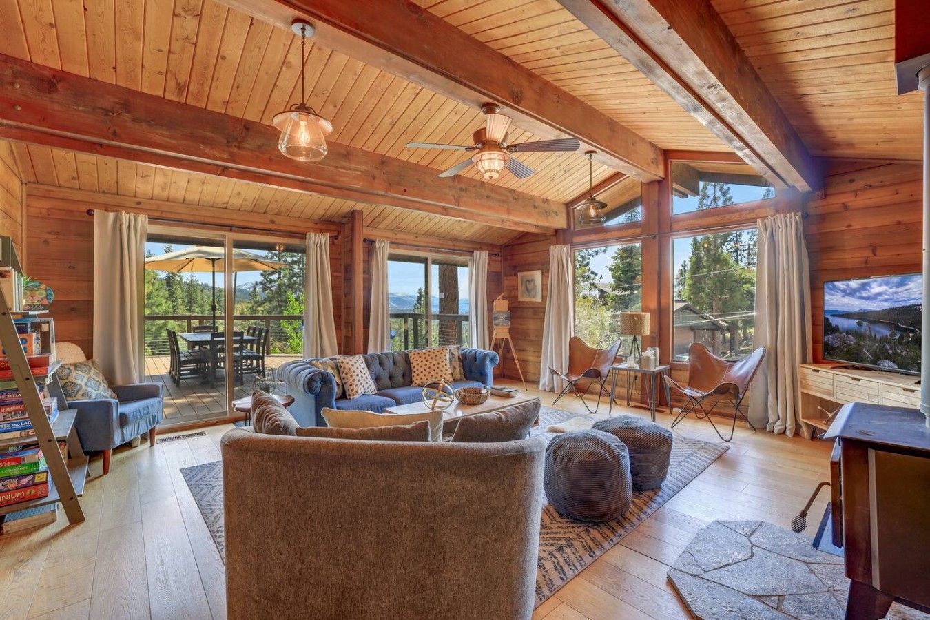 A spacious living room with a large sofa, a coffee table, and a television. There are multiple windows providing natural light and views of the outdoors.
