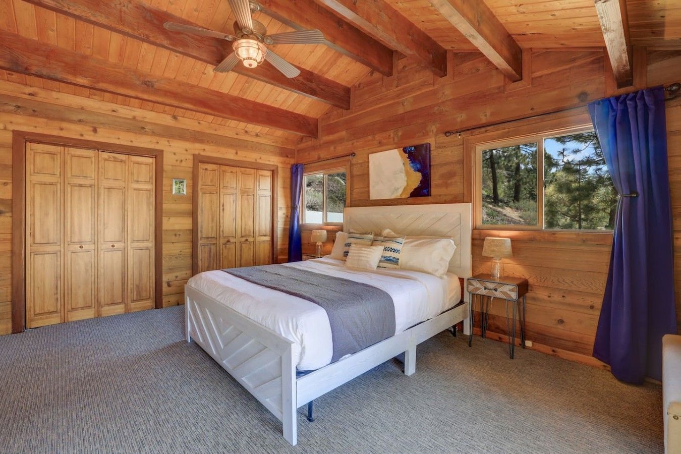Une chambre dans une cabane en bois, avec un lit, une table de chevet, un ventilateur de plafond et une grande fenêtre avec des rideaux.