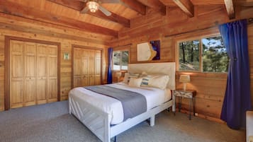 Une chambre dans une cabane en bois, avec un lit, une table de chevet, un ventilateur de plafond et une grande fenêtre avec des rideaux.