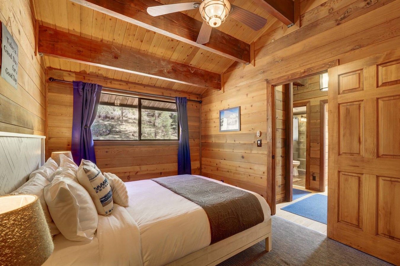 A wooden cabin bedroom with a bed, a ceiling fan, a window with curtains, and a door leading to a bathroom.