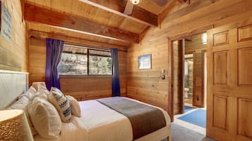 A wooden cabin bedroom with a bed, a ceiling fan, a window with curtains, and a door leading to a bathroom.