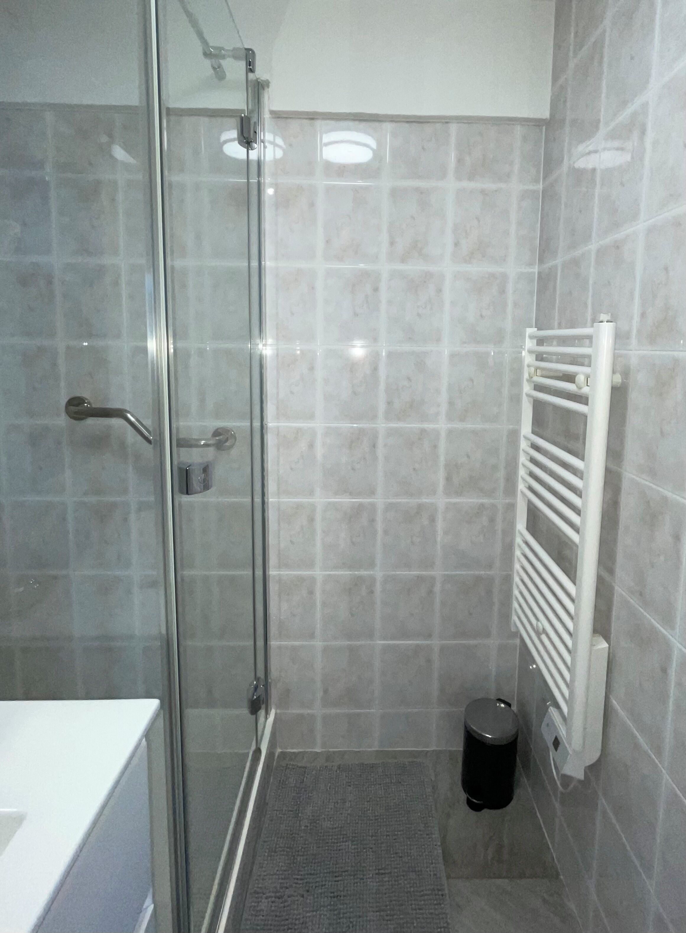 A modern bathroom with a glass shower enclosure, a white towel rack, and a small trash bin.