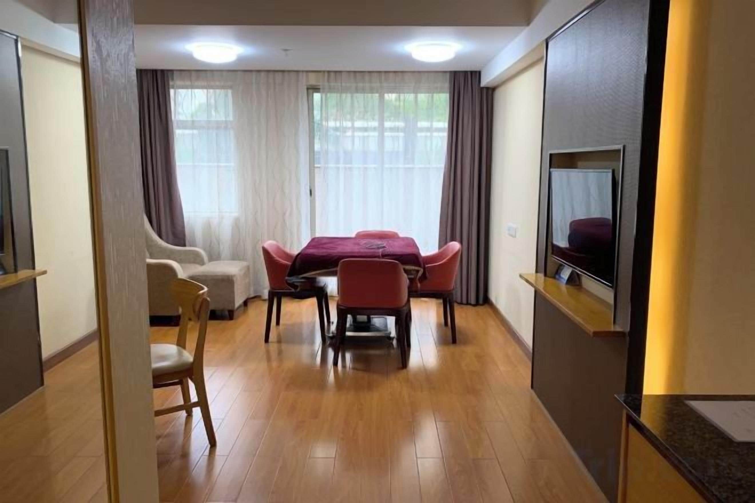 A hotel room with a wooden floor, a dining table with chairs, a sofa, a TV mounted on the wall, and a window with curtains.