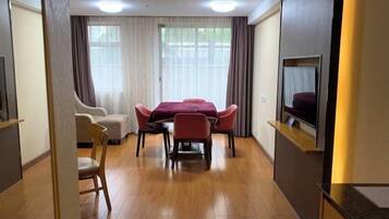 A hotel room with a wooden floor, a dining table with chairs, a sofa, a TV mounted on the wall, and a window with curtains.