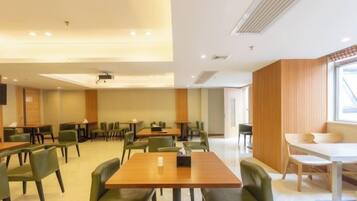 A spacious dining area with multiple tables and chairs, a projector screen, and a large window providing natural light.