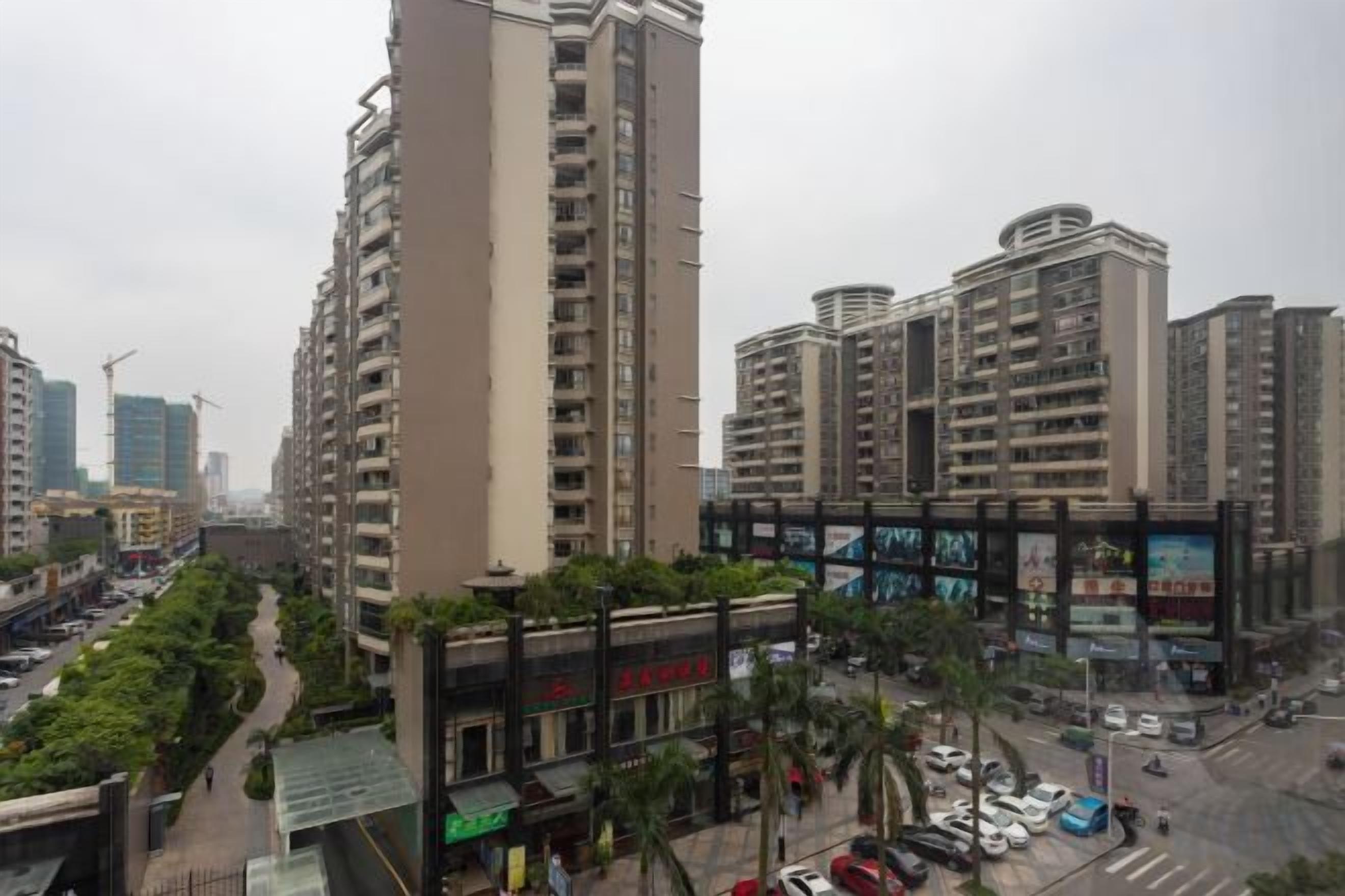 A high-rise residential building with multiple balconies, a pedestrian walkway, and a commercial area with shops and parked cars.