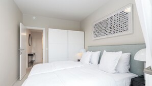 Une chambre moderne avec un grand lit, une table de chevet avec une lampe, une œuvre d’art accrochée au mur et une porte donnant sur une autre pièce.