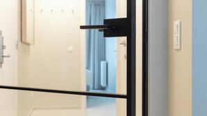 Une chambre d’hôtel moderne avec une porte en verre, un couloir visible et un interrupteur sur le mur.