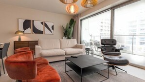 Un salon moderne avec un canapé, une table basse noire et un fauteuil rouge. Deux lampes suspendues sont placées au-dessus du canapé, et une grande fenêtre offre une vue sur les bâtiments environnants.