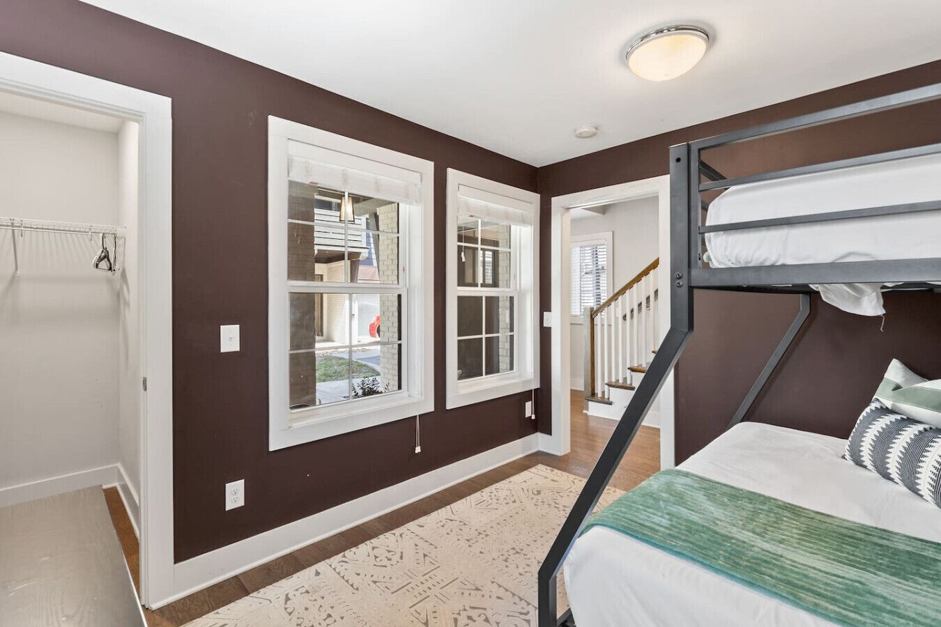 A bedroom with a bunk bed, two windows, a staircase, and a hallway with a coat rack.