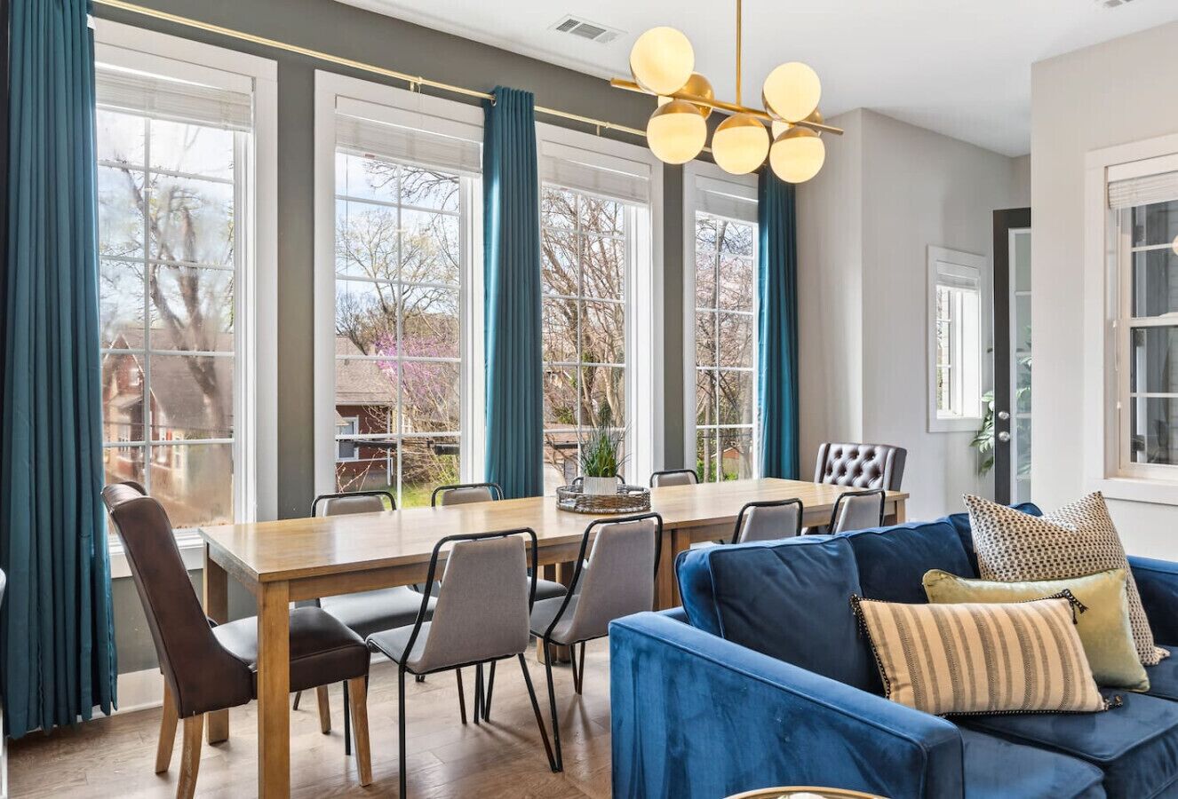 En moderne stue med en blå sofa, spisebord og stole. Store vinduer med gardiner giver naturligt lys og udsigt til omgivelserne.