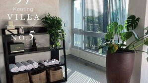 A modern living room with a large window, a black shelving unit with towels, a large potted plant, and a wooden bench. - 57villa (Hengchun)