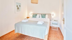 Ein ordentlich gemachtes Bett mit einer hellblauen Bettdecke, zwei Kissen mit Blättermotiv, ein Nachttisch mit Lampe und ein gerahmtes Bild an der Wand.