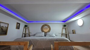 Un dormitorio con una cama, un espejo, dos mesitas de noche e iluminación azul en el techo.