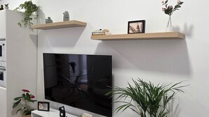 Una sala de estar moderna con un televisor de pantalla plana grande, muebles blancos, estanterías de madera y plantas decorativas.