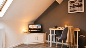 Una stanza accogliente con soffitto spiovente, una televisione su un mobile, un tavolo con una sedia e un quadro appeso al muro.