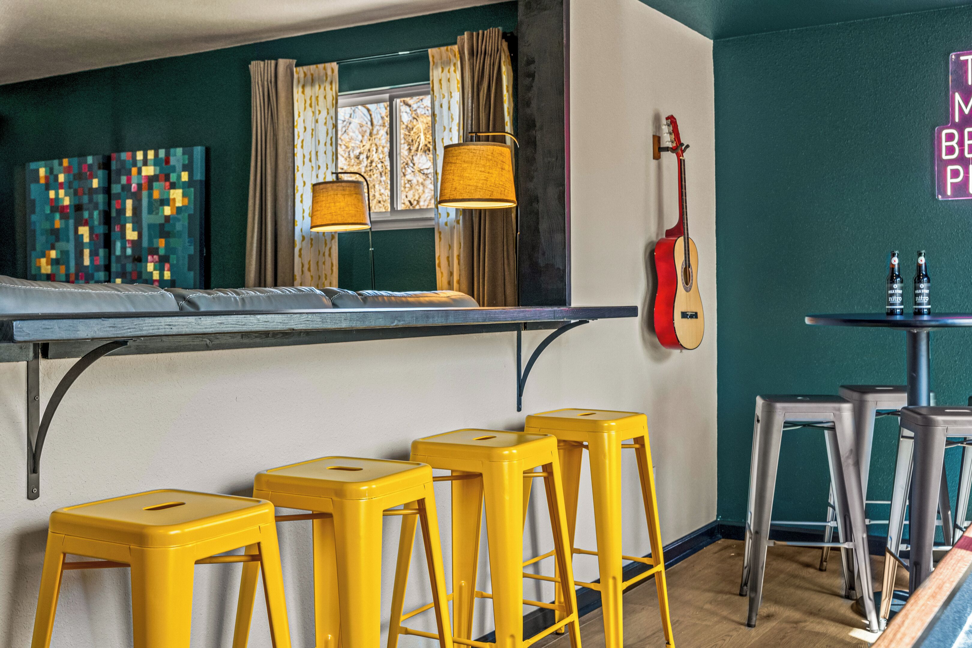 A room with a bed, yellow stools, a guitar, and a table with bottles.