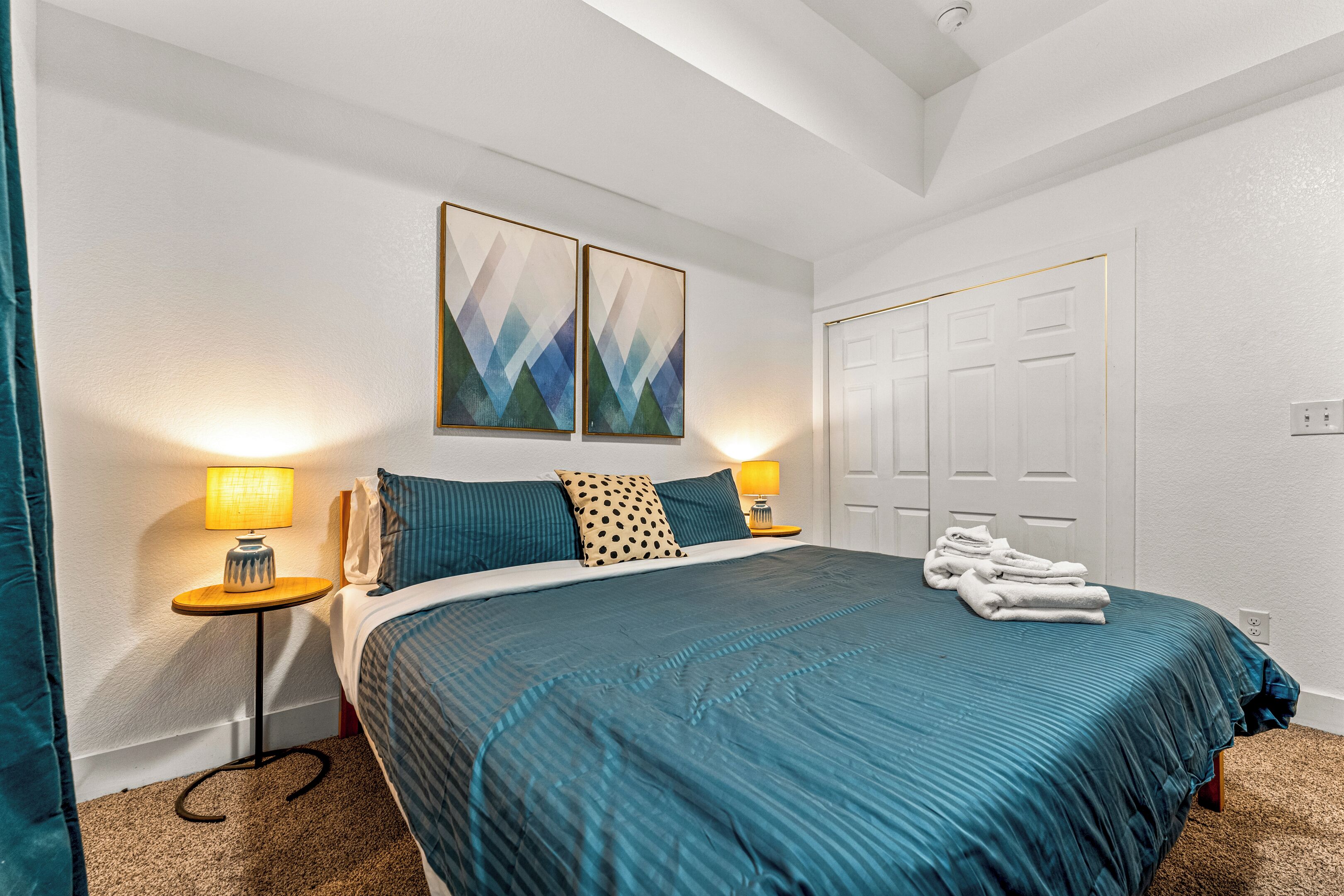 A neatly arranged bedroom with a bed, two wall-mounted lamps, a pair of abstract paintings, and a set of white doors.