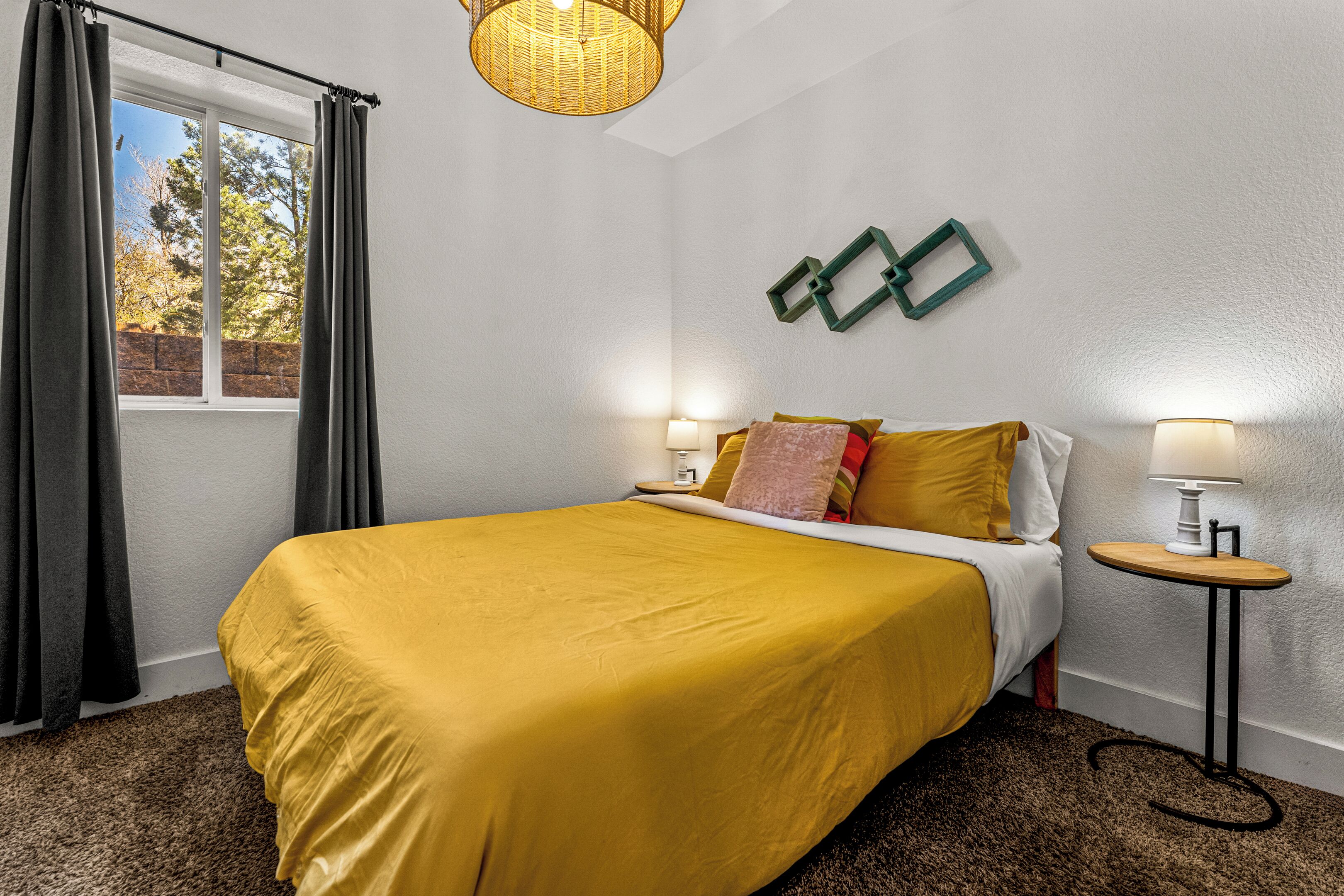 A bedroom with a large bed, a window with curtains, a bedside table with a lamp, and a wall-mounted light fixture.