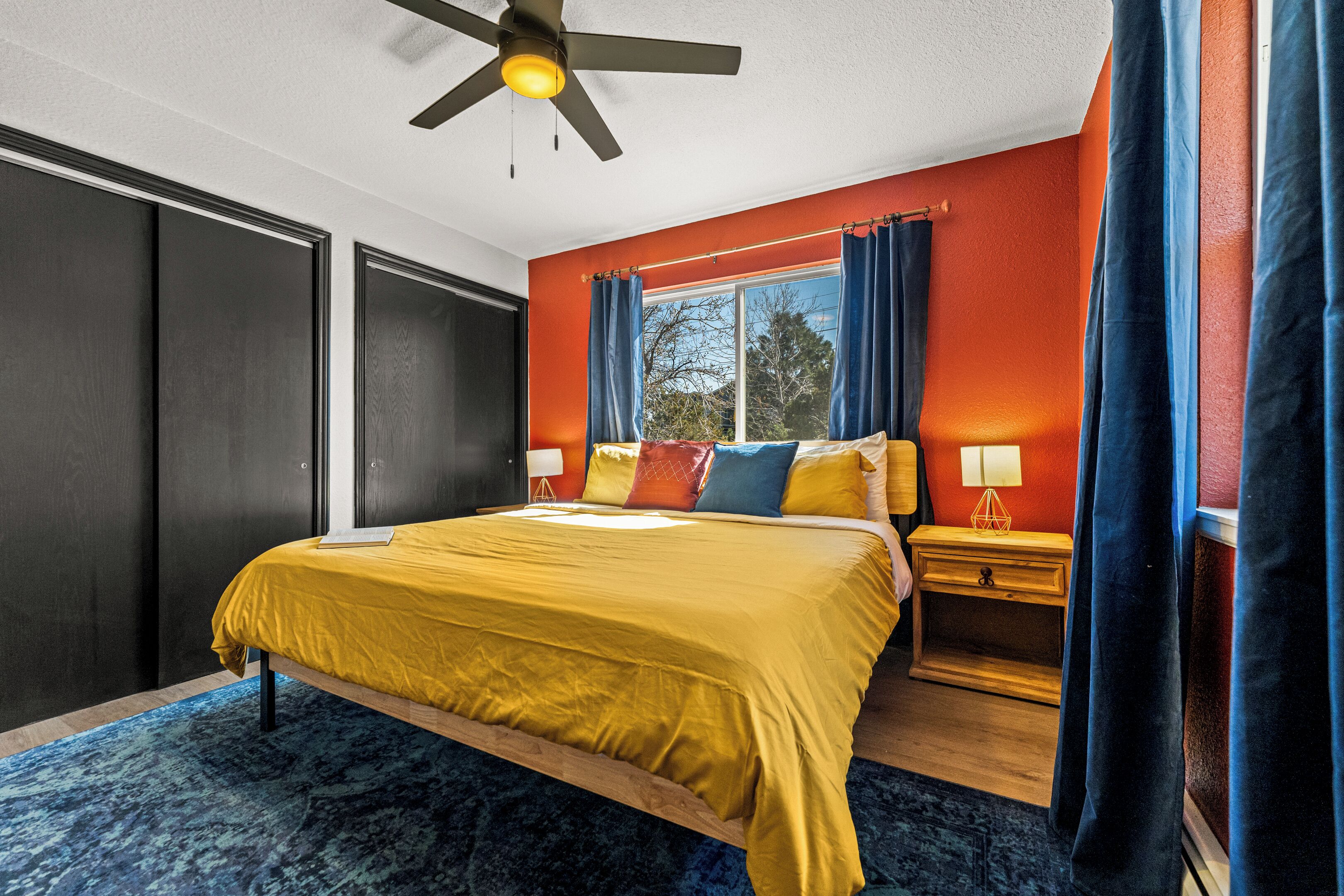 A bedroom with a bed, a nightstand, a window with curtains, a ceiling fan, and a view of trees outside.