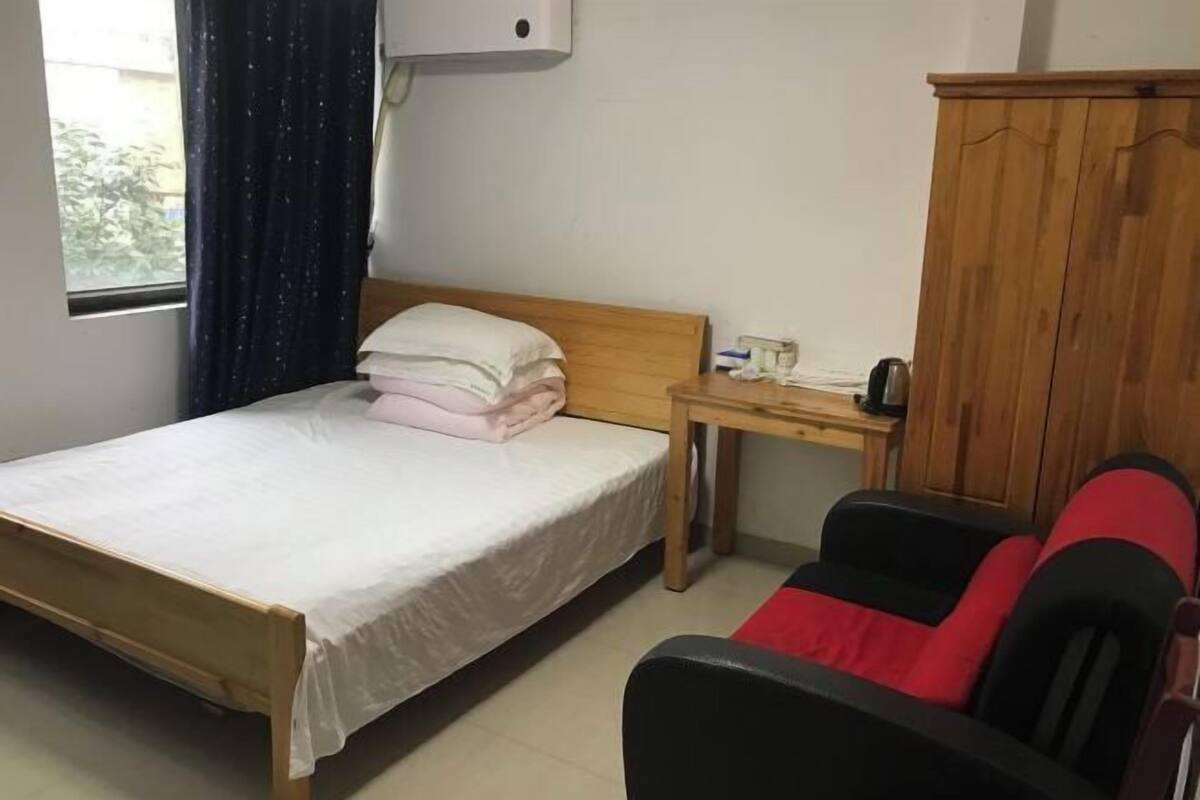 A single bed with a wooden headboard, a bedside table with a phone and a cup, a window with a curtain, and a wooden wardrobe.