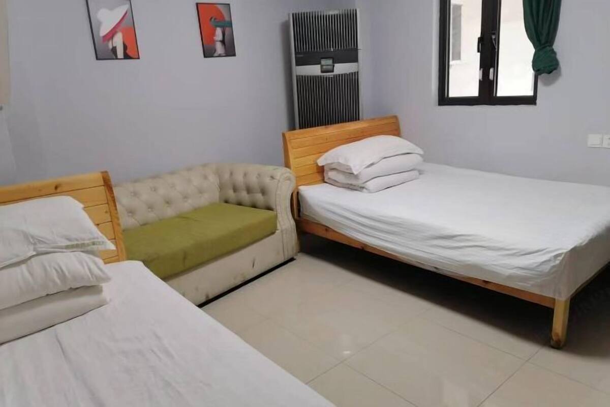 A room with two beds, a sofa, a wall-mounted air conditioner, and a window with curtains.