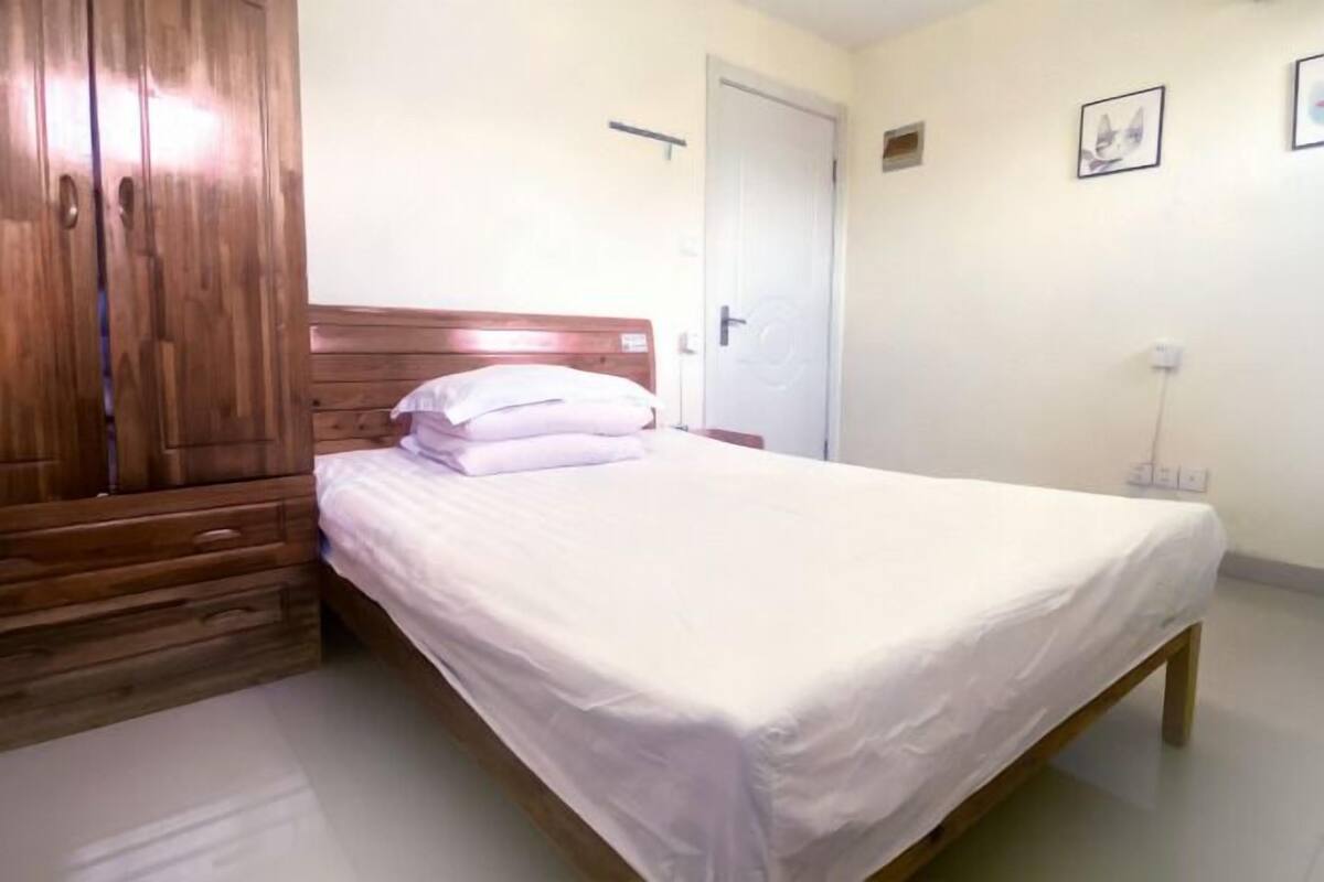 A bedroom with a wooden wardrobe, a bed with white bedding, a door, and a wall-mounted light fixture.