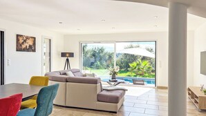 Een moderne woonkamer met een hoekbank, een eettafel en stoelen. Er is een groot raam met uitzicht op een zwembad en groen.