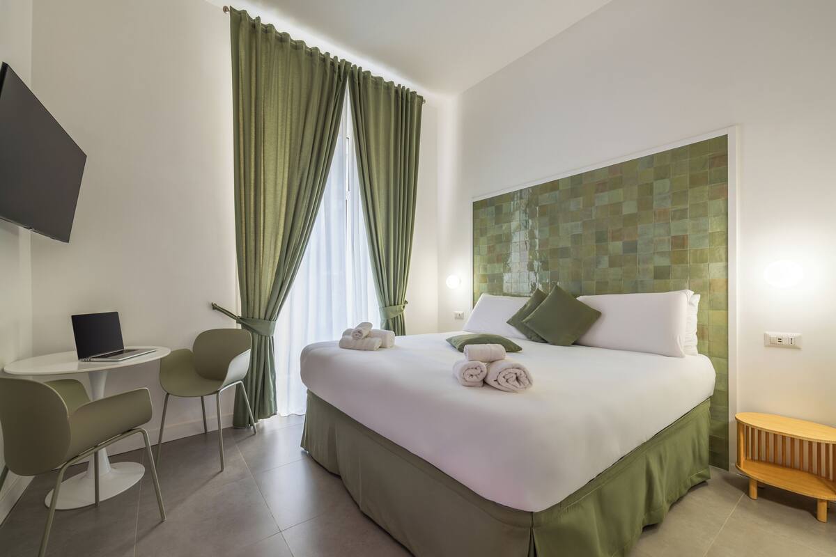 A hotel room with a bed, a desk with a laptop, two chairs, a TV, and a window with curtains.