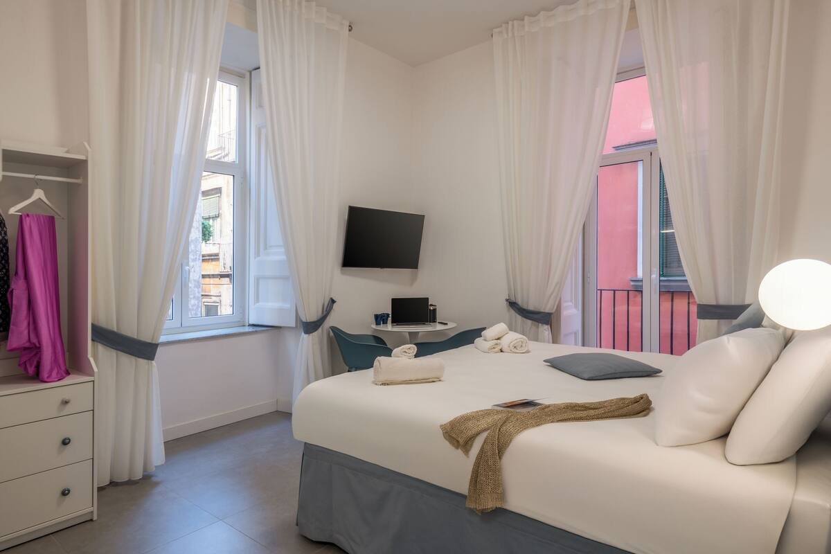 A bedroom with a bed, a TV, a bedside table, a window with curtains, a wardrobe, and a balcony view.