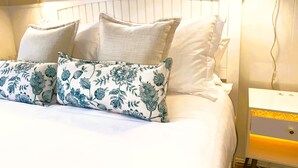 Una cama bien hecha con almohadas estampadas con motivos florales, cabecera blanca y mesita de noche con lámpara.