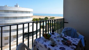 A balcony with a table set for dining, overlooking a beach and a white building. - Cozy studio with balcony and sea view (Bibione)