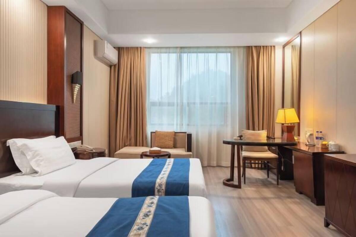Habitación de hotel con dos camas, un escritorio, una silla, una lámpara y un ventanal grande con cortinas.