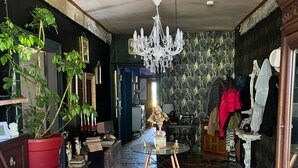 Een kamer met een kroonluchter, potplanten, een tafel met een vaas, en een etalagepop.
