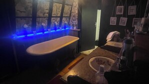 Een gedimd verlichte badkamer met een bad, een wastafel en een spiegel. Er hangen foto's in lijsten aan de muur en een lamp boven de spiegel.