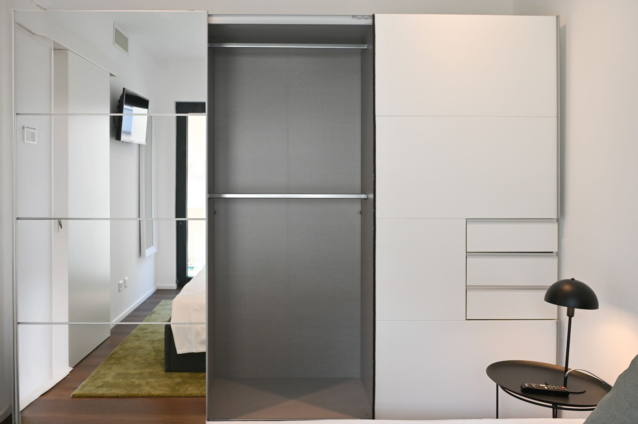 A modern hotel room with a sliding door closet, a bed, a small table with a lamp, and a TV mounted on the wall.