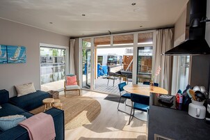 Una sala de estar moderna con sofá, mesa de centro y sillas, una zona de cocina con estufa y alacenas, y un puerta corrediza de vidrio que da a una terraza exterior con mesa y sillas.